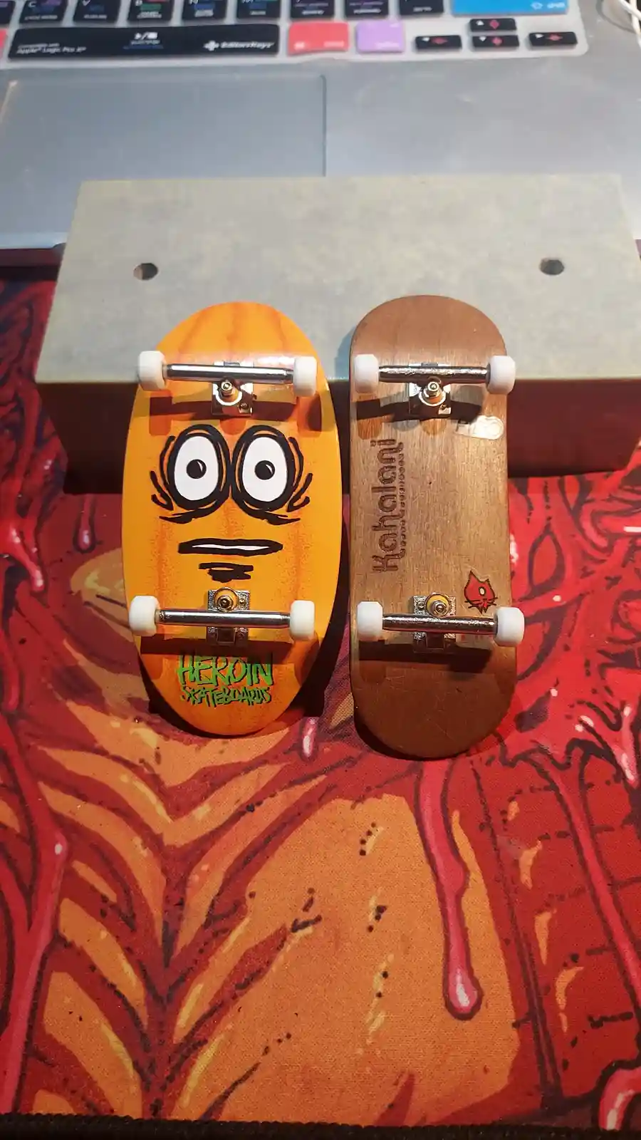 Two miniature finger skateboards sit side-by-side on a laptop keyboard, one orange with a cartoon face and 'HEROIN…