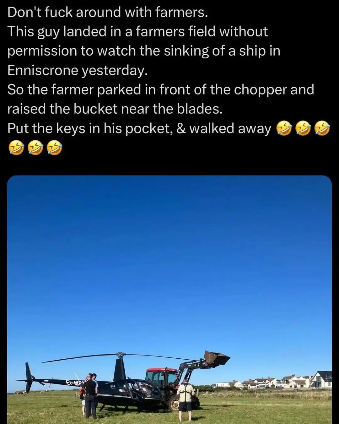 A black helicopter is parked on a grassy field under a clear blue sky, with a red tractor parked directly in front of it…