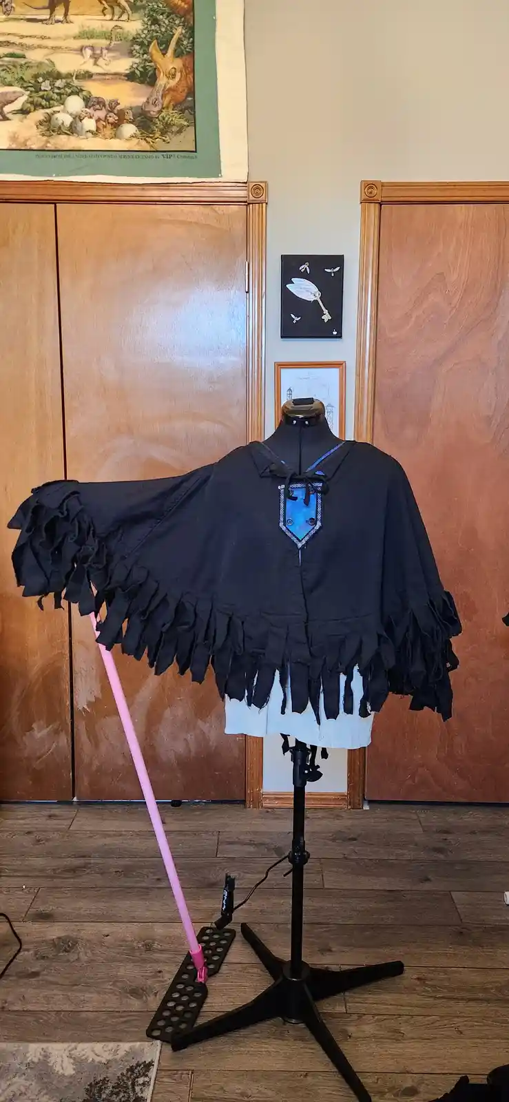 A homemade black cape with ruffled edges and a blue shield emblem is displayed on a mannequin stand. A pink mop handle is…