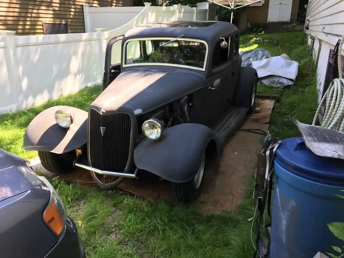 A matte black vintage hot rod, likely a 1930s Ford, sits in a grassy backyard on a wooden plank, its engine bay exposed and…