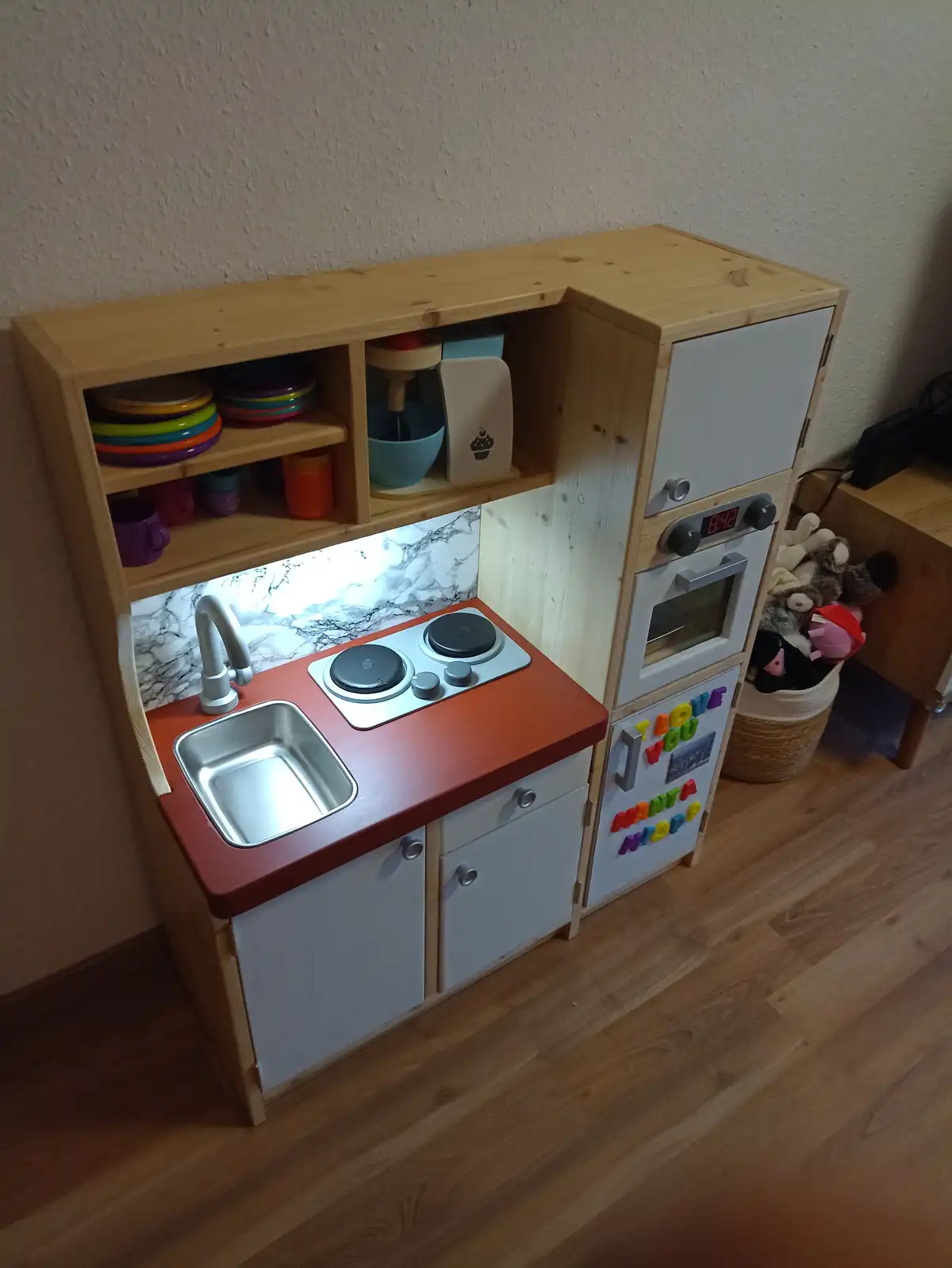 A wooden children's play kitchen with a red countertop, sink, stove, and oven, featuring colorful toy dishes on open shelves…