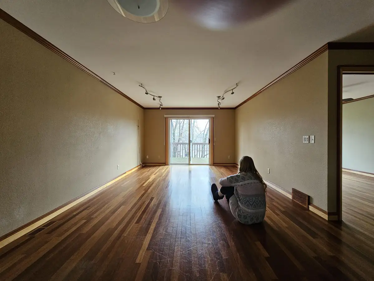 A person sits alone on the hardwood floor of an empty, unfurnished apartment, facing a glass door that leads outside. The…