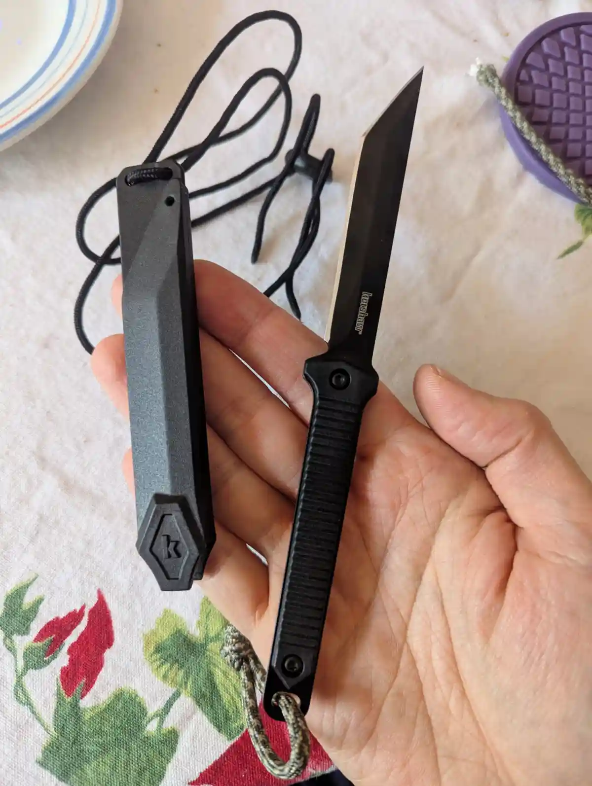 A hand holds a black Kershaw knife next to its matching black sheath, both featuring the brand's 'k' logo, on a…
