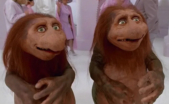 Two ape-like creatures with long brown hair and wide eyes stand in a brightly lit, sterile-looking room with people in the…