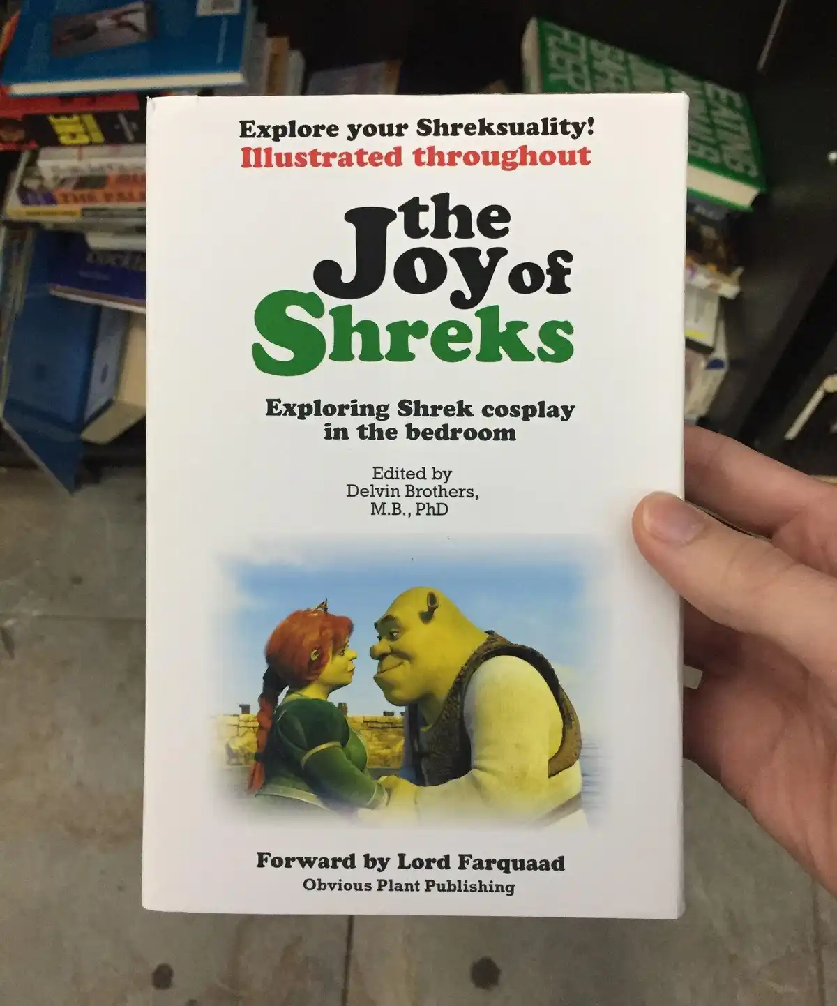 A hand holds a mock book cover parodying 'The Joy of Sex' with 'The Joy of Shreks' and 'Exploring Shrek cosplay in the…
