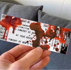 A hand holds a concert ticket that has been heavily splattered with red paint or fake blood, obscuring much of the text. The…