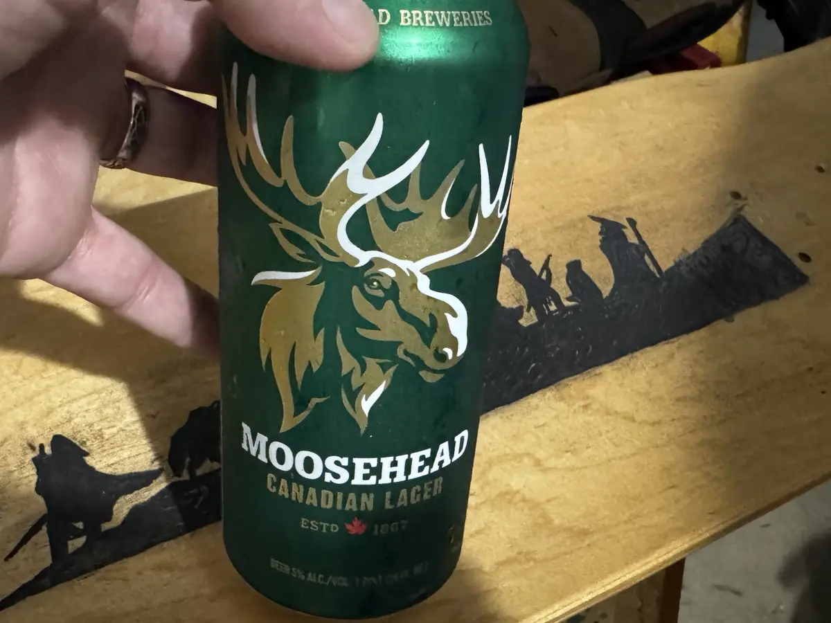 A hand holds a green Moosehead Canadian Lager beer can, featuring a large moose head logo, against a wooden surface with…