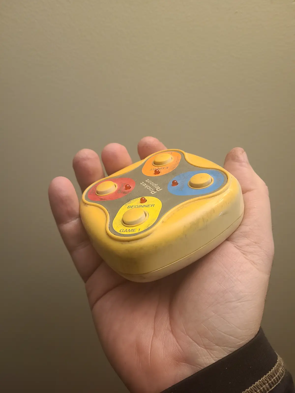 A hand holds a yellow, worn, retro handheld game console with four colored buttons labeled 'BEGINNER', 'AMATEUR', 'EXPERT'…