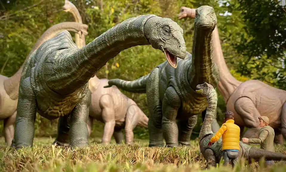 A photo shows several large, realistic-looking dinosaur toys in a grassy outdoor setting, with two small human figurines…