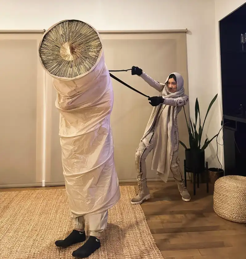 A person in a beige, textured costume with a hood and gloves is pulling on a black strap attached to a large, cylindrical…