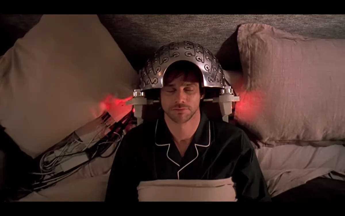 a man in black pajamas lies in bed with his eyes closed, wearing a futuristic silver helmet with red glowing lights on…