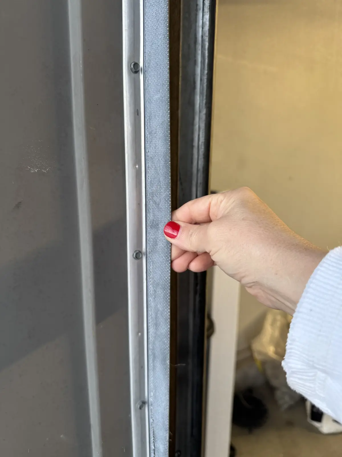 A close-up photo shows a person's hand with red-painted fingernails gripping a narrow, textured grey door seal strip between…