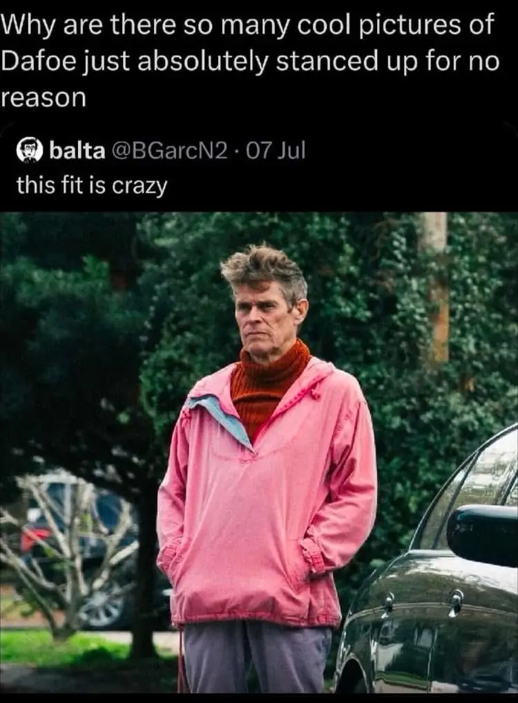 A man with messy hair, wearing a pink jacket over a rust-colored turtleneck and gray pants, stands with his hands in his…