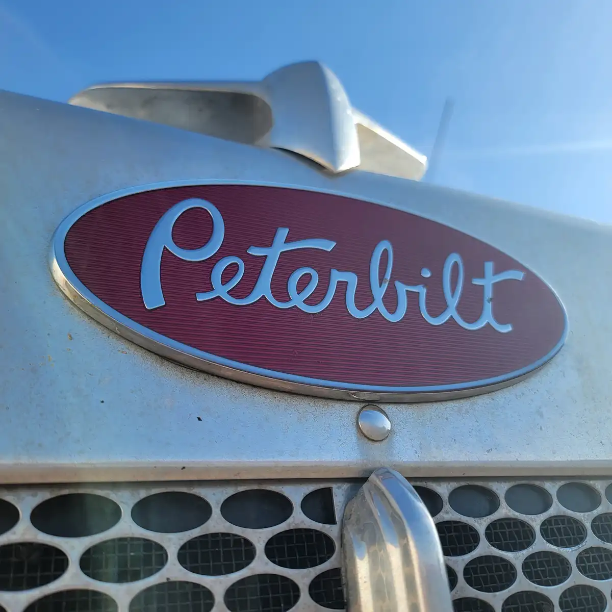 A close-up photo shows the chrome 'Peterbilt' logo on a vintage truck's grille, set against a clear blue sky. The oval…