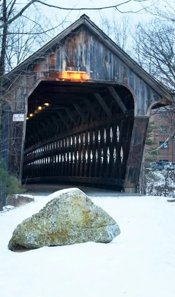 A snowy scene featuring a large, mossy rock in the foreground, with a wooden covered bridge in the background. The bridge…