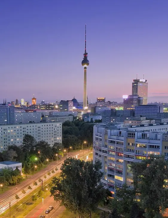 A dusk photograph of Berlin's skyline featuring the iconic Berlin TV Tower rising above residential buildings and lit…