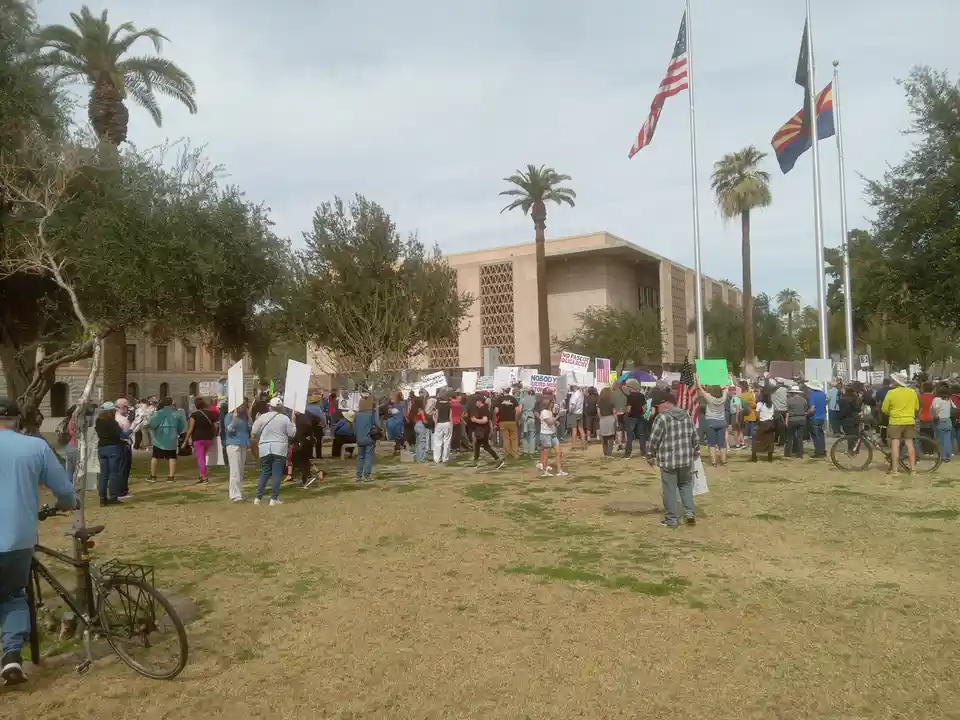 A large outdoor protest is visible with many people holding signs in front of a modern building, under American and Arizona…