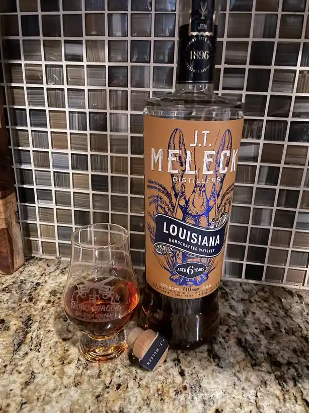 A bottle of J.T. Meleck Distillers Louisiana Handcrafted Whiskey, aged 6 years, sits next to a partially filled whiskey…