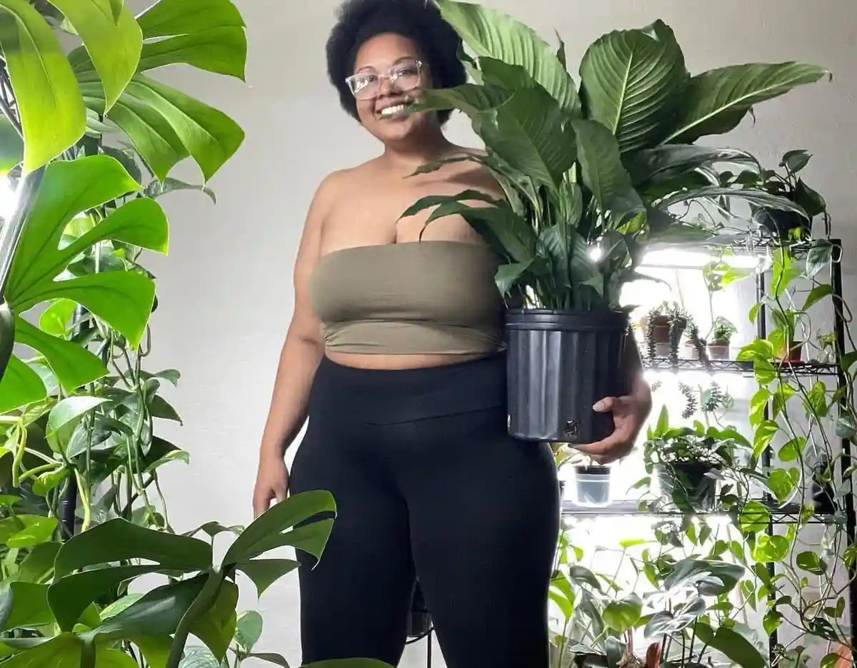 A smiling woman with glasses and an afro stands surrounded by lush green houseplants, holding a large potted plant. She…