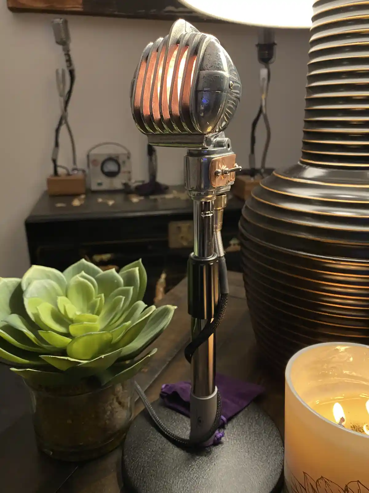 A vintage-style microphone-shaped lamp with a glowing bulb inside its grille sits on a table next to a potted succulent and…