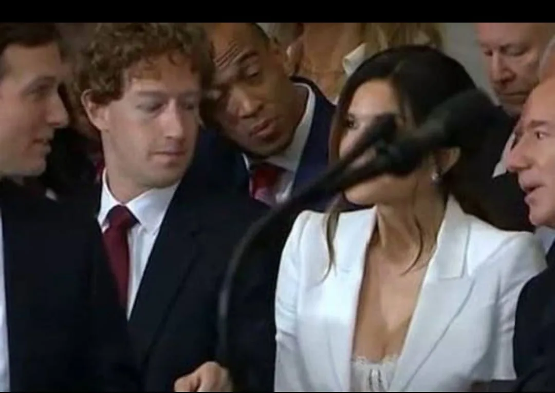 A woman in a white blazer speaks into a microphone while surrounded by several men in suits, including Mark Zuckerberg and…