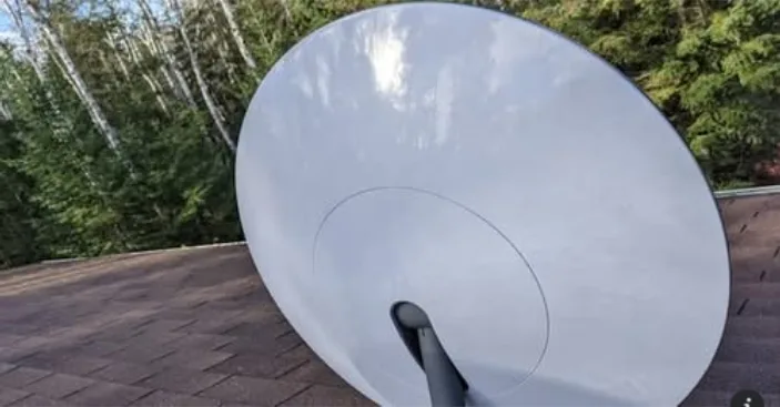 A large white satellite dish is mounted on a brown shingled roof, angled slightly upward, with trees and sky visible in the…