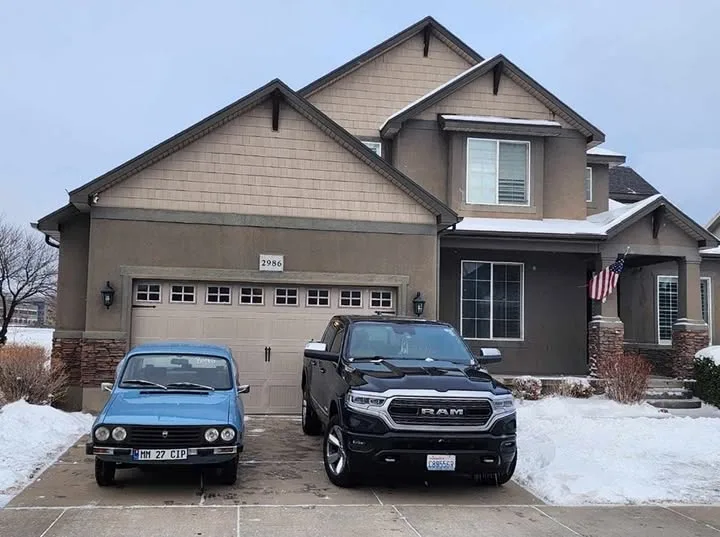 A snowy suburban driveway shows a large, modern two-story house with a garage. Parked in front are two contrasting vehicles…
