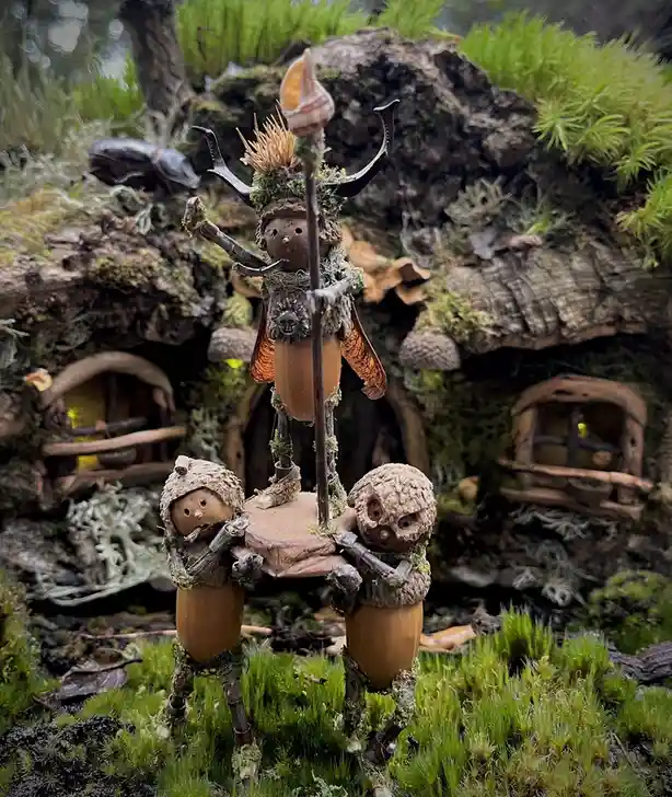 Three whimsical, handcrafted figurines made from acorns, twigs, and moss stand in a mossy forest diorama. The central…