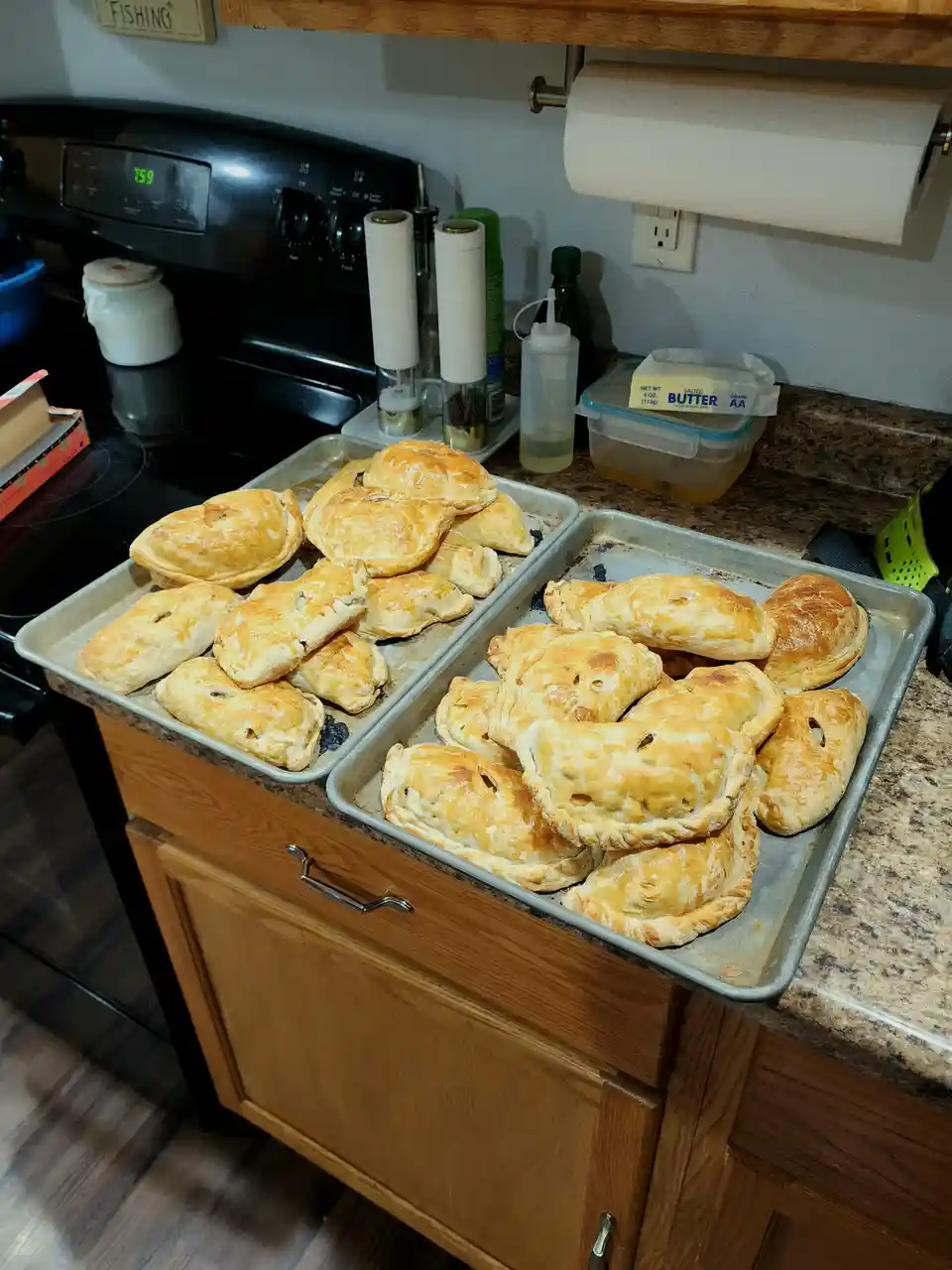 Two metal baking trays filled with golden-brown, freshly baked empanadas sit on a granite kitchen counter next to a stove…