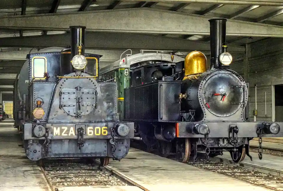 Two vintage steam locomotives are parked side-by-side on tracks inside a large industrial-style shed. The locomotive on the…