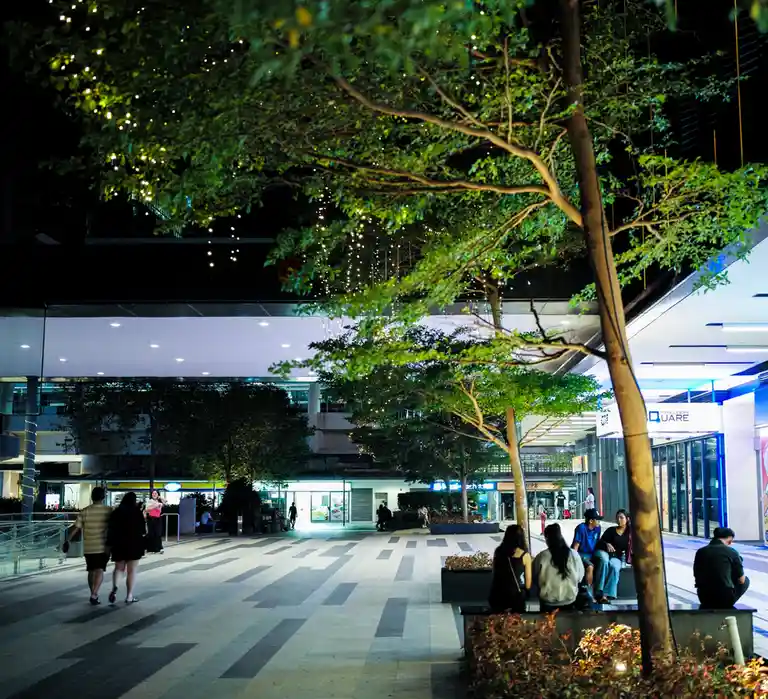 A nighttime photo of a modern outdoor shopping plaza with people walking and sitting on benches under trees decorated with…