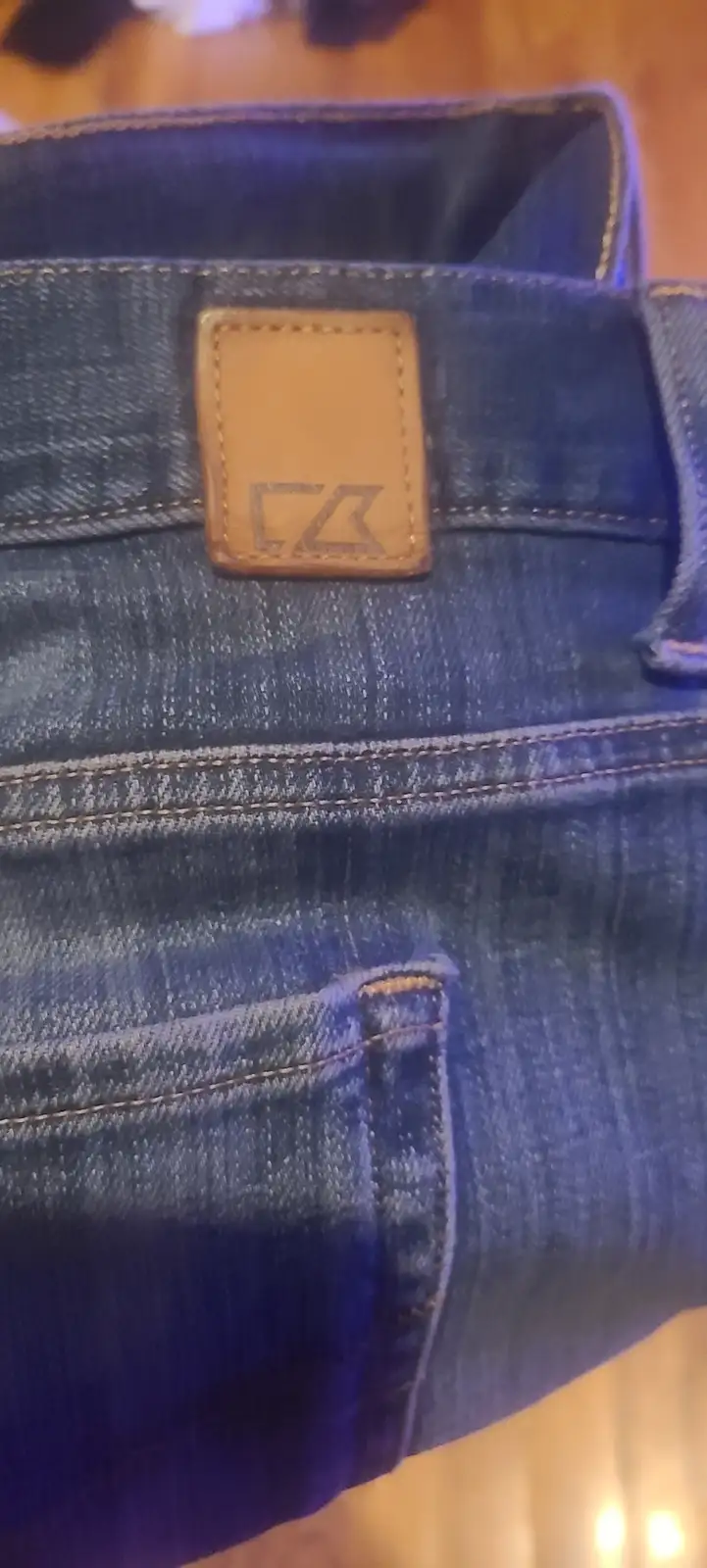 A close-up photo of blue denim jeans showing a brown leather brand patch with a stylized 'CZ' logo, positioned above a back…