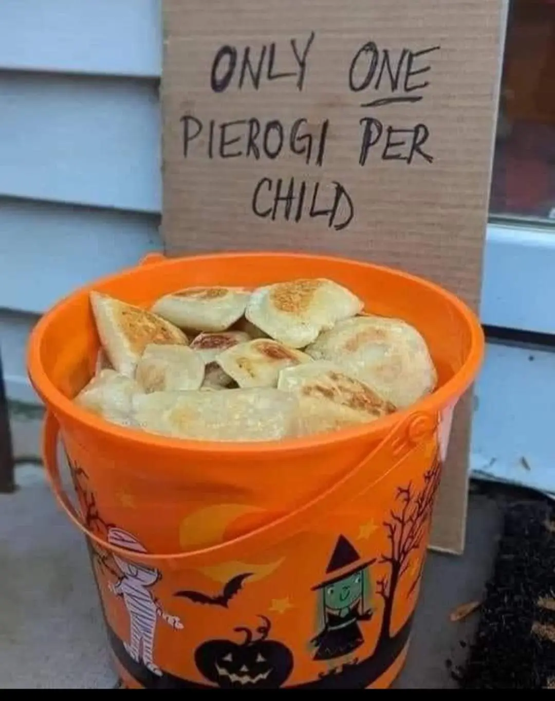 An orange Halloween-themed bucket filled with fried pierogi sits on a porch, with a handwritten cardboard sign behind it…