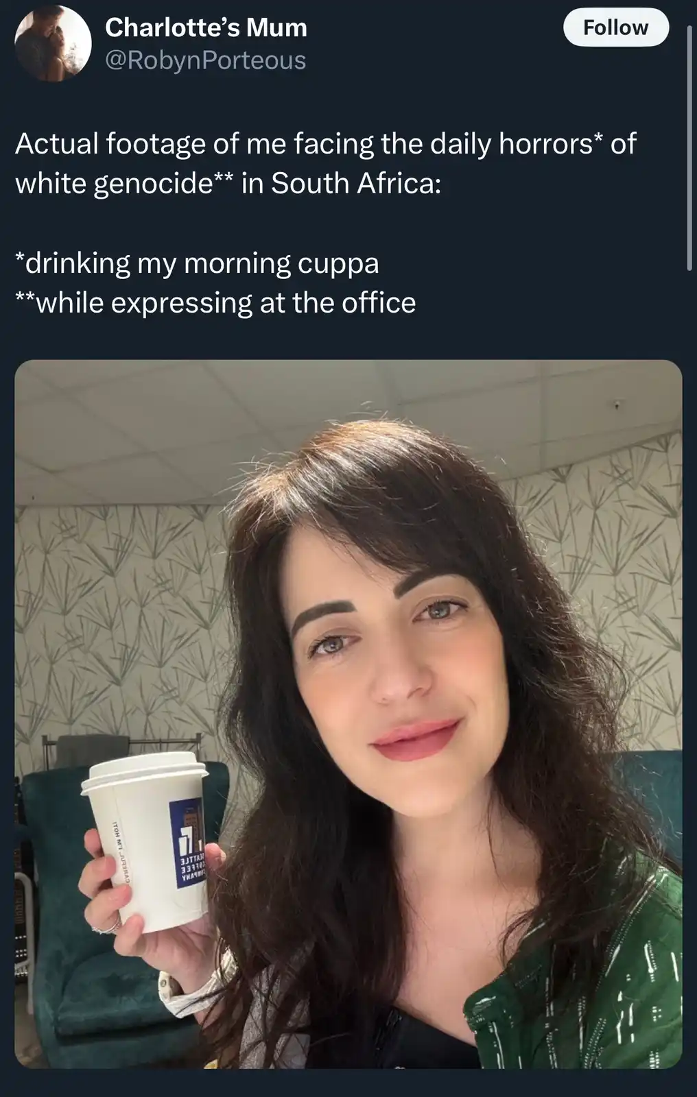 A woman with dark hair and a green jacket holds a white coffee cup while smiling at the camera in an office setting with…