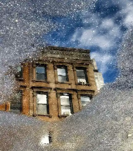A puddle on asphalt reflects a brownstone building and a blue sky with clouds, distorted by the water's surface. The image…