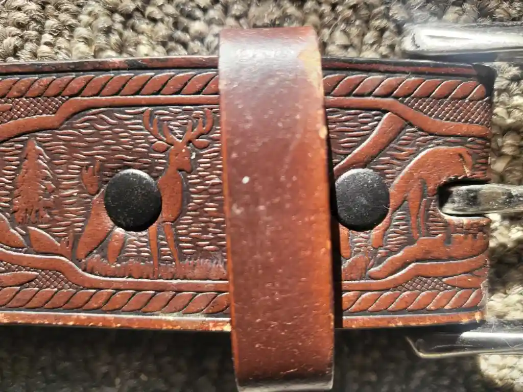 A close-up of a brown leather belt buckle with embossed designs, showing a deer on the left side and a dog on the right…