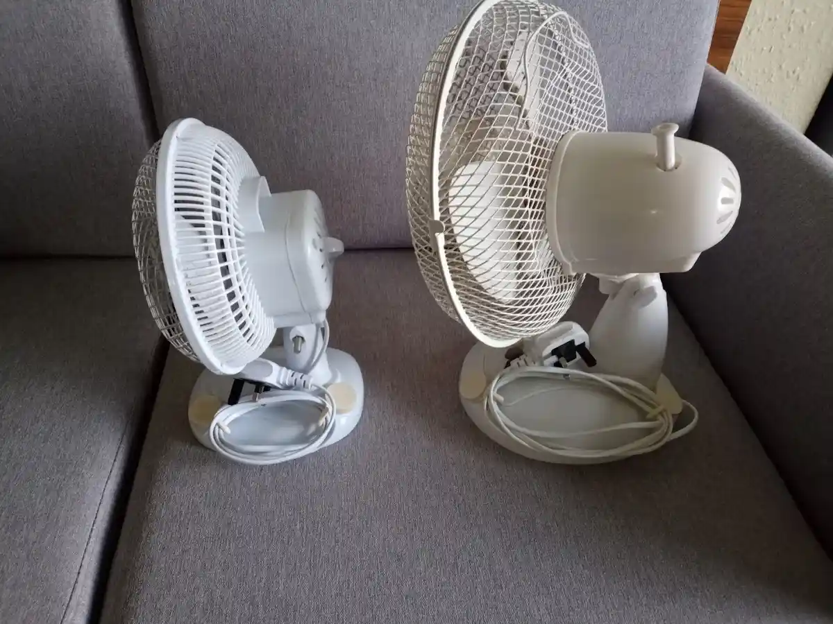Two white electric fans sit facing each other on a gray fabric couch, their cords coiled neatly at their bases. The overlaid…