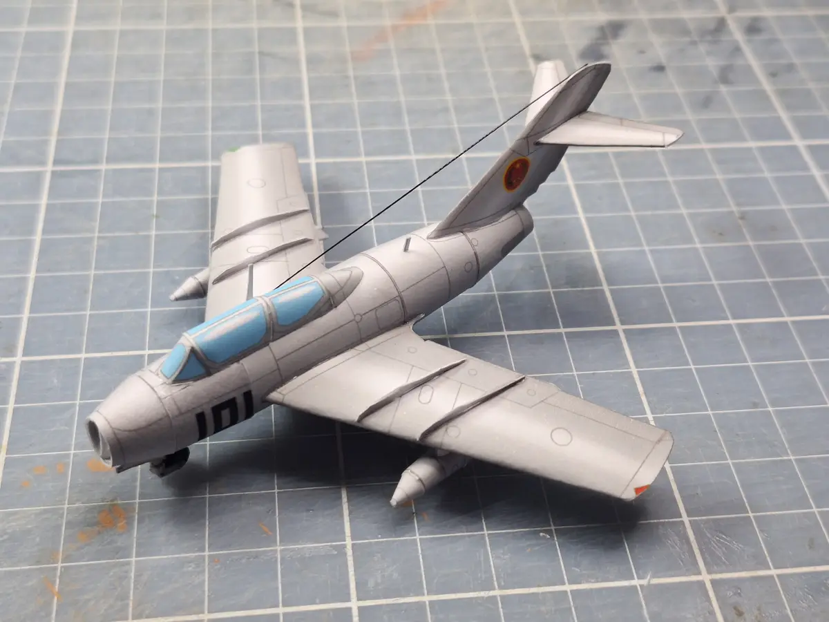 A handmade paper airplane model, resembling a military jet with gray body, blue cockpit, and Chinese roundel on the tail…