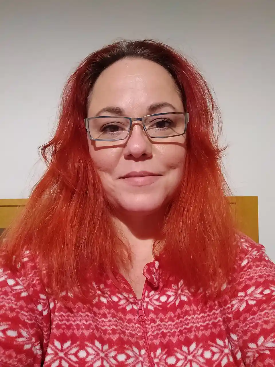 A woman with long, vibrant red hair and glasses wears a red and white patterned zip-up fleece top while seated. The overlaid…