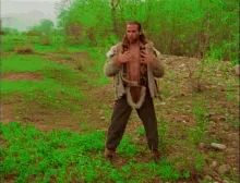 A man in a rugged, open jacket stands in a grassy field, initially shirtless then later with a brick wall in front of him…