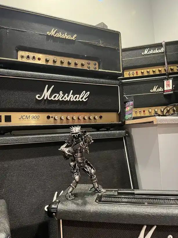 A silver, intricate metal sculpture of a humanoid figure playing a guitar stands atop a black Marshall guitar amplifier…