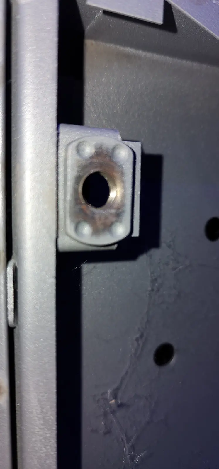 A close-up photo shows a metal door hinge with four bolts securing it to a metal door frame. The hinge has a central…