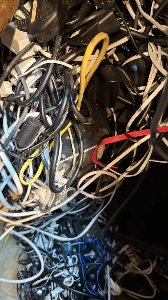 A close-up photo shows a chaotic tangle of various electrical cords, plugs, and adapters in black, white, yellow, and blue…