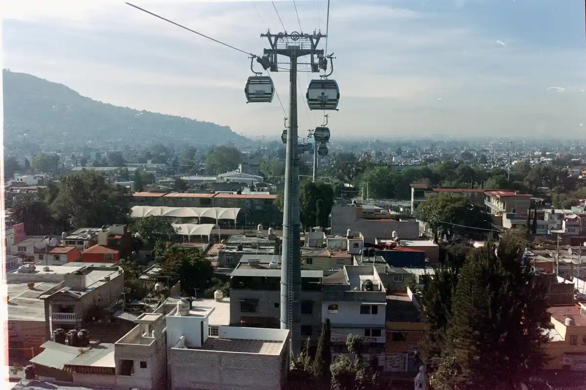 An aerial view of a cityscape with a prominent cable car system, featuring two gondolas suspended from overhead cables…