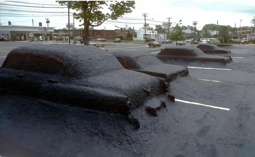 A row of vintage cars in a parking lot are completely covered in thick, black tar, with only their outlines and shapes…