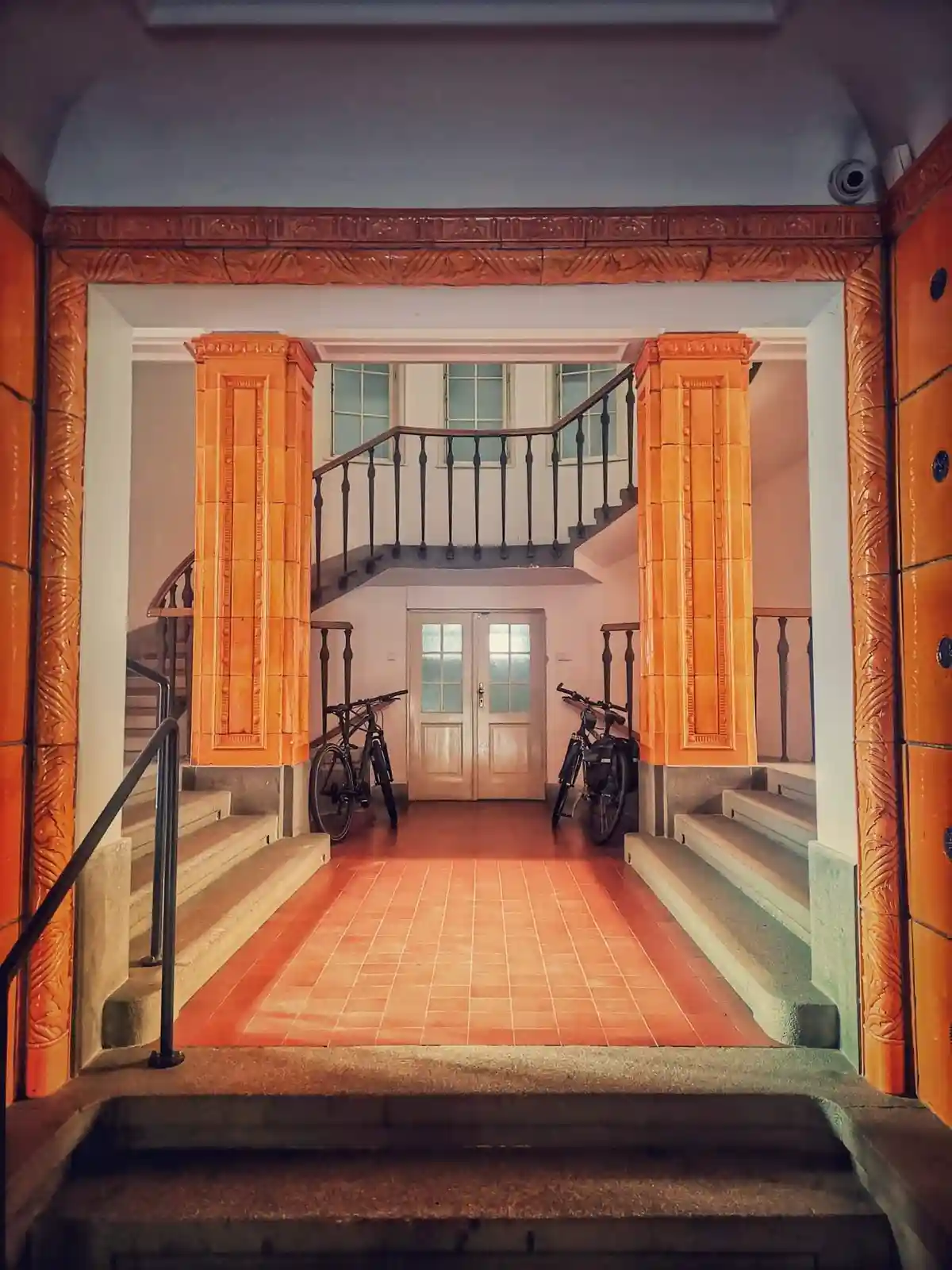 An ornate, tiled hallway with stairs on both sides leads to a landing with two bicycles parked beside a door. The walls and…