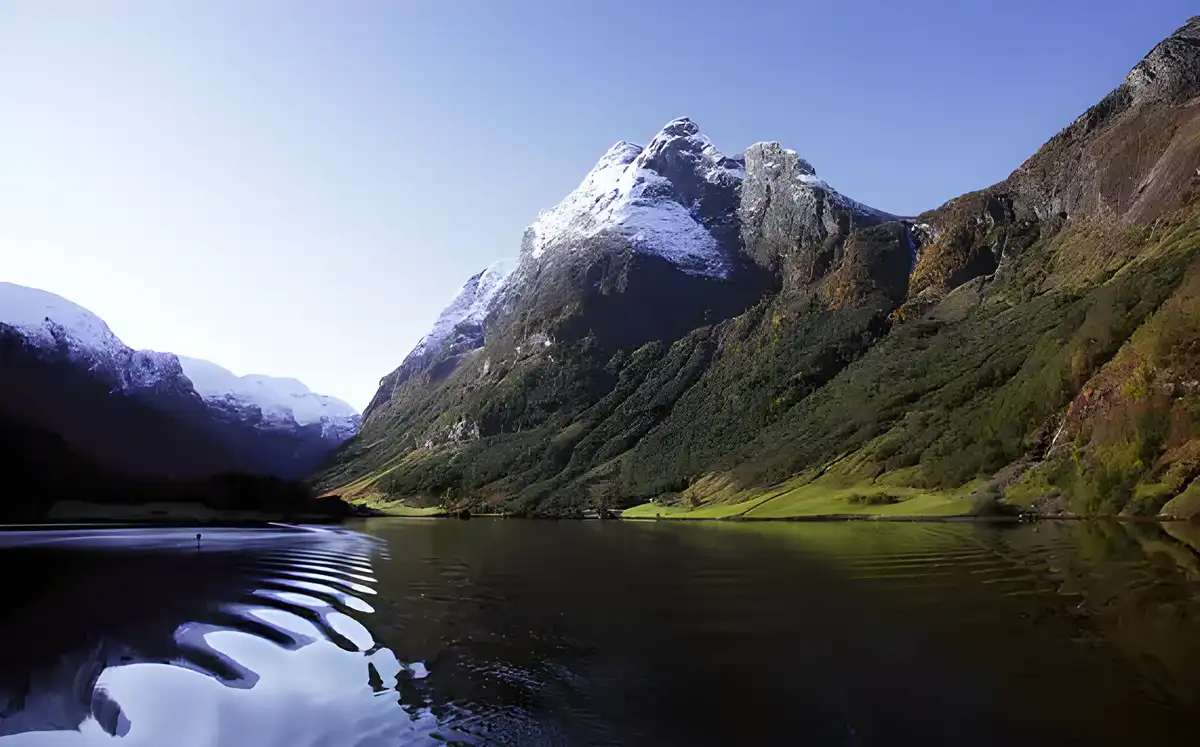 A serene landscape photo shows a calm lake reflecting snow-capped mountains under a clear blue sky, with lush green slopes…