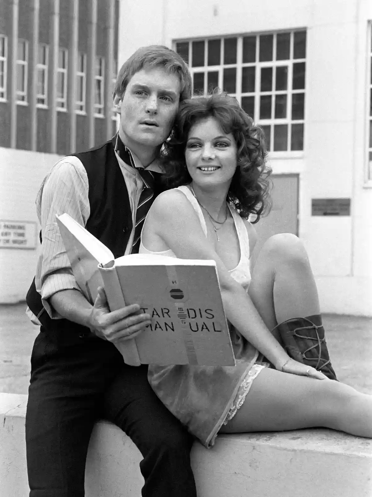 A black-and-white photo shows a man and woman sitting on a ledge outdoors. The man, wearing a vest and tie, holds an open…