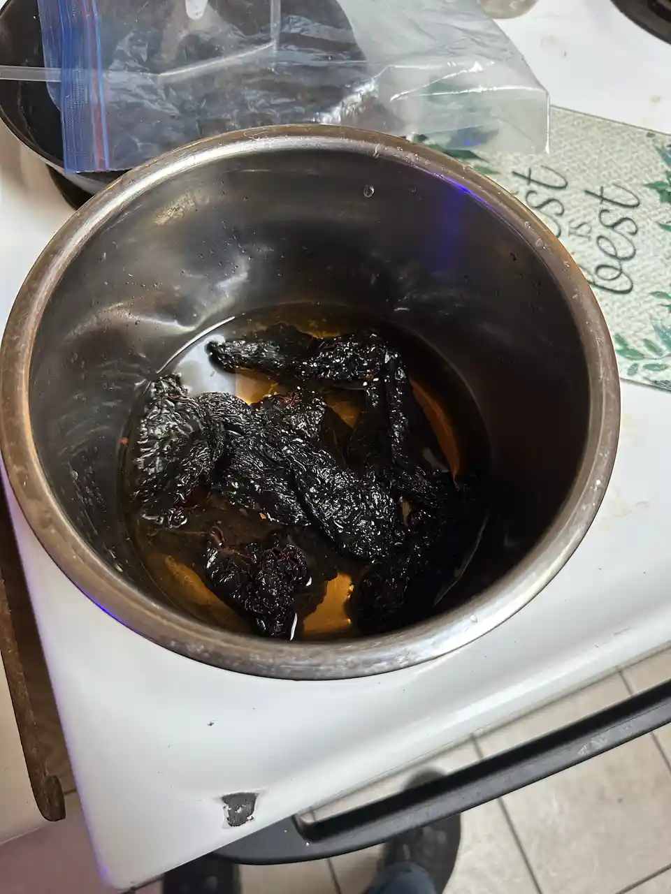 A stainless steel bowl sits on a white stovetop, containing several very dark, shriveled, and charred-looking dried chiles…