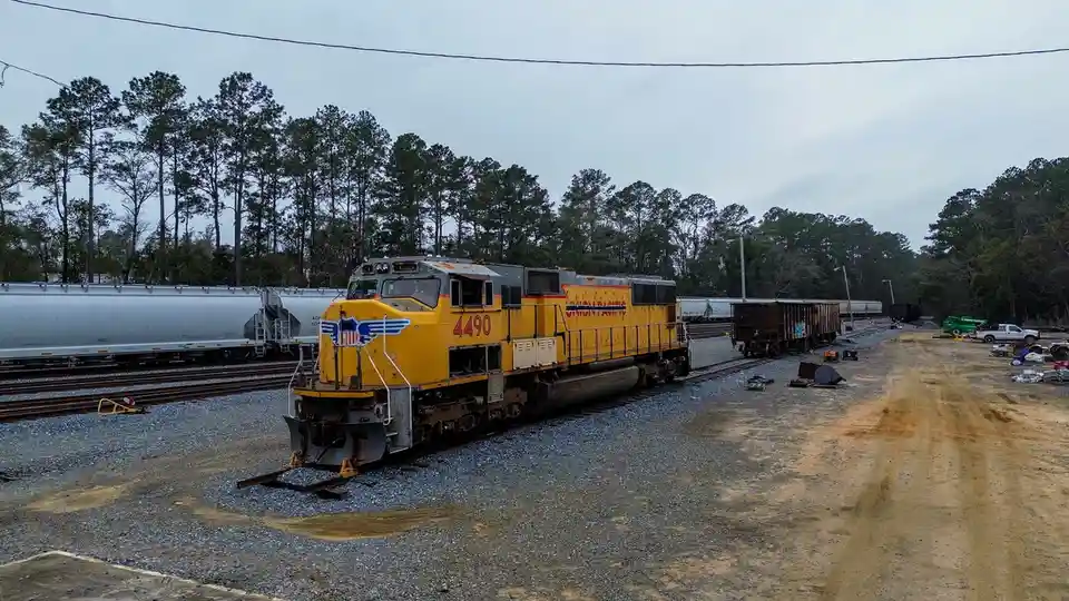 A yellow Union Pacific locomotive, numbered 4490, sits on tracks in a rail yard surrounded by trees and other railcars under…